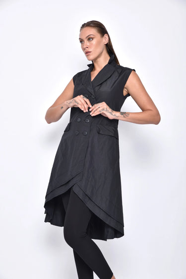 A woman wearing a sleeveless black trench-style Vest with a double-breasted button front, notched lapel collar and flap pockets on either side of the waist, asymmetrical hem line, paired with black leggings and chunky black open-toe heels over a white background.