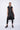 A woman wearing a sleeveless black trench-style Vest with a double-breasted button front, notched lapel collar and flap pockets on either side of the waist, asymmetrical hem line, paired with black leggings and chunky black open-toe heels over a white background.