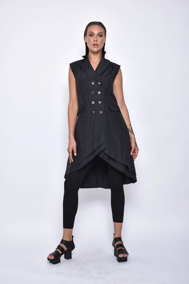 A woman wearing a sleeveless black trench-style Vest with a double-breasted button front, notched lapel collar and flap pockets on either side of the waist, asymmetrical hem line, paired with black leggings and chunky black open-toe heels over a white background.