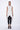 A woman wearing a sleeveless light grey trench-style Vest with a double-breasted button front, notched lapel collar and flap pockets on either side of the waist, asymmetrical hem line, paired with black leggings and chunky black open-toe heels over a white background.