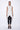 A woman wearing a sleeveless light grey trench-style Vest with a double-breasted button front, notched lapel collar and flap pockets on either side of the waist, asymmetrical hem line, paired with black leggings and chunky black open-toe heels over a white background.