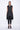 A woman wearing a sleeveless black trench-style Vest with a double-breasted button front, notched lapel collar and flap pockets on either side of the waist, asymmetrical hem line, paired with black leggings and chunky black open-toe heels over a white background.