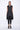 A woman wearing a sleeveless black trench-style Vest with a double-breasted button front, notched lapel collar and flap pockets on either side of the waist, asymmetrical hem line, paired with black leggings and chunky black open-toe heels over a white background.