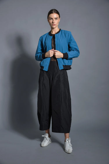 A woman wearing a blue bomber jacket and black pants against a gray background