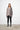 A woman standing against a white wall, wearing a 2 tone gray long-sleeve sweater with asymmetrical hem and black leggings.