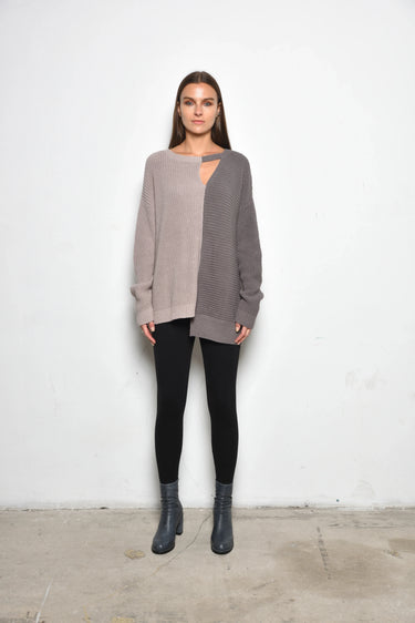A woman standing against a white wall, wearing a 2 tone gray long-sleeve sweater with asymmetrical hem and black leggings.