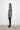 A woman standing against a white wall, wearing a 2 tone gray long-sleeve sweater with asymmetrical hem and black leggings.
