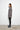 A woman standing against a white wall, wearing a 2 tone gray long-sleeve sweater with asymmetrical hem and black leggings.
