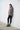 A woman standing against a white wall, wearing a gray long-sleeve sweater and black pants, with her feet positioned slightly apart.