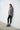 A woman standing against a white wall, wearing a gray long-sleeve sweater and black pants, with her feet positioned slightly apart.