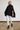 A woman modeling a black chunky knit poncho sweater with a high ribbed turtleneck, paired with off-white trousers and black leather ankle boots, standing against a white brick wall.