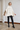 A woman modeling a cream cable-knit turtleneck poncho sweater, paired with black pants and black leather ankle boots, standing in a bright, white-walled room.