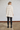 A woman viewed from the back wearing a cream-colored, textured knit turtleneck poncho sweater, black pants, and black boots, standing against a white brick wall.