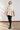 A woman modeling a chunky knit cream-colored poncho sweater, paired with black pants and black leather boots, standing in front of a white brick wall.