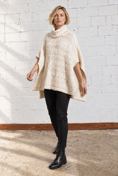 A woman modeling a chunky knit cream-colored poncho sweater, paired with black pants and black leather boots, standing in front of a white brick wall.