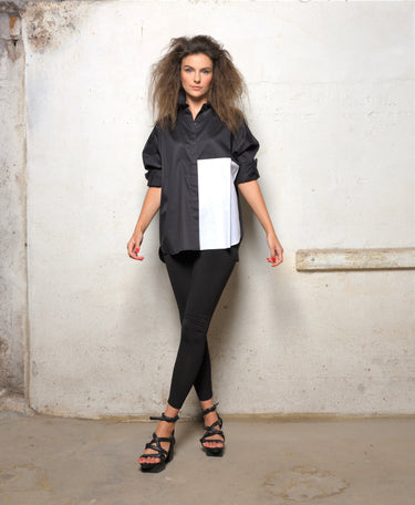 A woman modeling a 2 tone black and white boxy shirt with dolman sleeves, paired with black leggings and strappy sandals, standing against a textured wall.