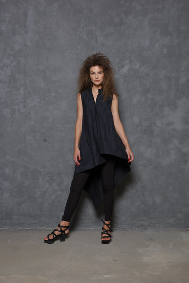 A woman modeling a sleeveless asymmetrical tunic shirt in black, paired with black pants and black strappy sandals, standing against a gray background.