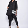 A woman wearing a black asymmetrical tunic shirt with a polka dot band design, paired with black leggings and black sandals over a white background.