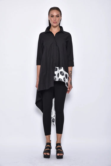 A woman wearing a black asymmetrical tunic shirt with a polka dot band design, paired with black leggings and black sandals over a white background.