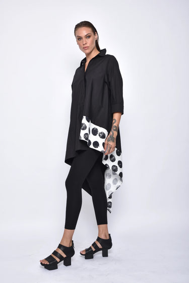 A woman wearing a black asymmetrical tunic shirt with a polka dot band design, paired with black leggings and black sandals over a white background.