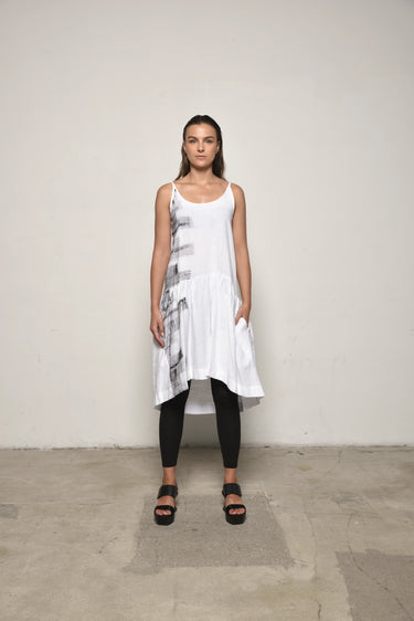 A female model wearing a spaghetti straps white peplum dress with strokes paint design on the side, pockets, low neck line, asymmetrical hem, paired with Black leggings, black sandals against an industrial white background.