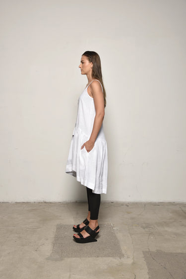 A female model wearing a spaghetti straps white peplum dress with strokes paint design on the side, pockets, low neck line, asymmetrical hem, paired with Black leggings, black sandals against an industrial white background.