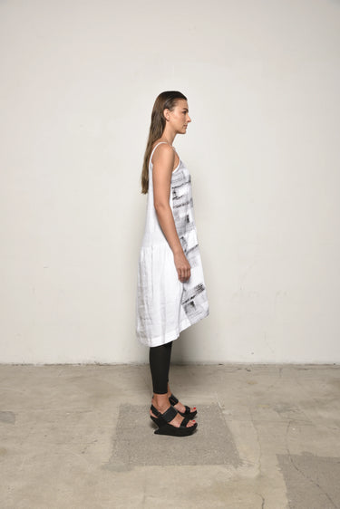 A female model wearing a spaghetti straps white peplum dress with strokes paint design on the side, pockets, low neck line, asymmetrical hem, paired with Black leggings, black sandals against an industrial white background.