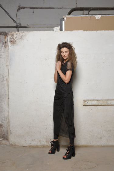 A woman standing in front of a white textured wall wearing a black mesh dress with short sleeves, a round neckline, and a maxi length. She is also wearing black high heels.