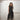 A woman standing in front of a white textured wall wearing a black mesh dress with short sleeves, a round neckline, and a maxi length. She is also wearing black high heels.