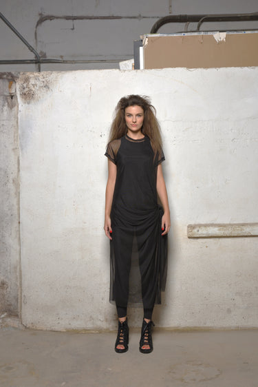 A woman standing in front of a white textured wall wearing a black mesh dress with short sleeves, a round neckline, and a maxi length. She is also wearing black high heels.