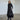 A model wearing a long black dress with long sleeves, cowl neck an A-line shape, high-low hem, pockets and a concealed zipper on the side.
