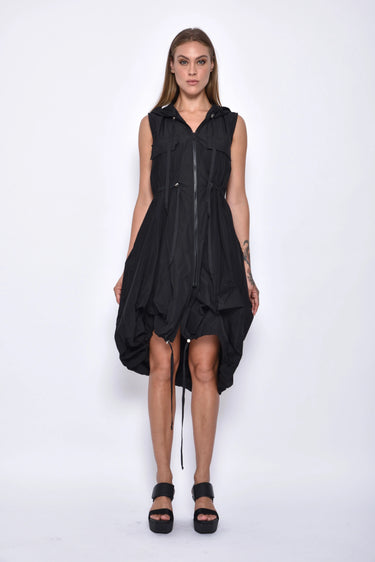 A woman wearing a sleeveless black dress with a sporty, utilitarian design. The dress includes a hood, a front zipper closure, and drawstring details at the waist and hem, allowing for adjustable ruching. The silhouette is voluminous and sculptural, particularly around the hemline, which forms a balloon-like shape, chunky black platform sandals. The background is plain white.