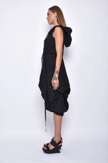A woman wearing a sleeveless black dress with a sporty, utilitarian design. The dress includes a hood, a front zipper closure, and drawstring details at the waist and hem, allowing for adjustable ruching. The silhouette is voluminous and sculptural, particularly around the hemline, which forms a balloon-like shape, chunky black platform sandals. The background is plain white.