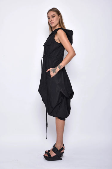 A woman wearing a sleeveless black dress with a sporty, utilitarian design. The dress includes a hood, a front zipper closure, and drawstring details at the waist and hem, allowing for adjustable ruching. The silhouette is voluminous and sculptural, particularly around the hemline, which forms a balloon-like shape, chunky black platform sandals. The background is plain white.