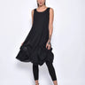 A  woman wearing a sleeveless black sheer dress with a scoop neckline. The dress has a voluminous, sculptural silhouette with gathered and ruched detailing around the hemline, paired with black leggings and chunky black open-toe heels sandals over a white background.