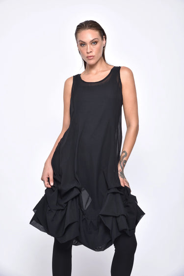 A  woman wearing a sleeveless black sheer dress with a scoop neckline. The dress has a voluminous, sculptural silhouette with gathered and ruched detailing around the hemline, paired with black leggings and chunky black open-toe heels sandals over a white background.