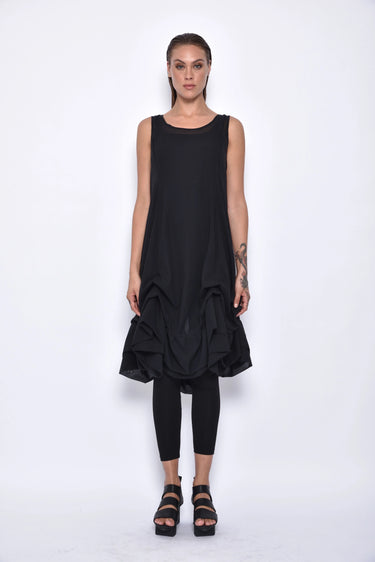 A  woman wearing a sleeveless black sheer dress with a scoop neckline. The dress has a voluminous, sculptural silhouette with gathered and ruched detailing around the hemline, paired with black leggings and chunky black open-toe heels sandals over a white background.
