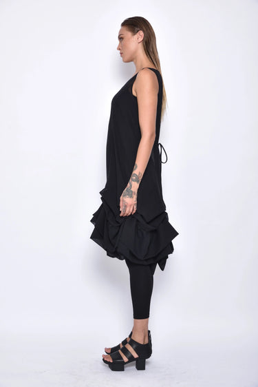 A  woman wearing a sleeveless black sheer dress with a scoop neckline. The dress has a voluminous, sculptural silhouette with gathered and ruched detailing around the hemline, paired with black leggings and chunky black open-toe heels sandals over a white background.
