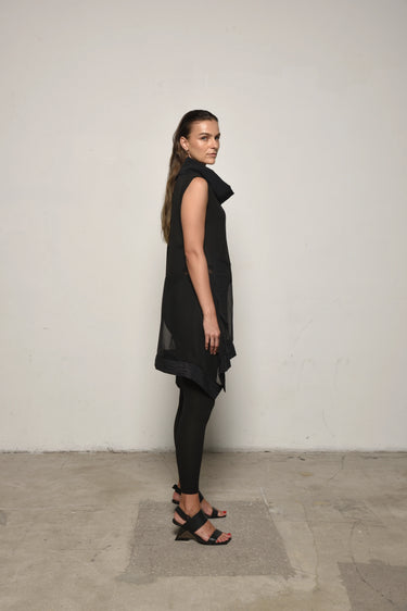 A woman modeling a sleeveless black tunic with a draped cowl neck and Asymmetrical hem, paired with black leggings.