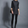 A woman modeling a black top with 3/4 sleeves and textured pattern, paired with black leggings and black boots against a gray background