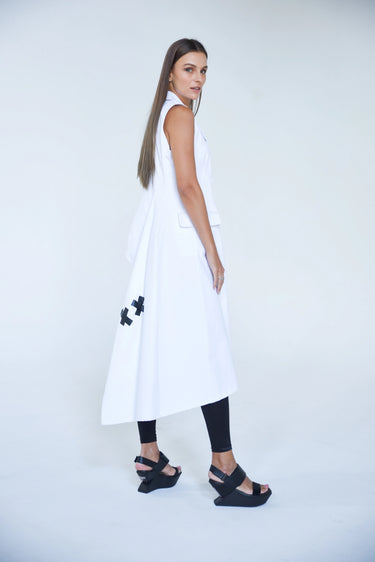 A woman standing against a white backdrop wearing an asymmetrical sleeveless White vest and Black leggings.