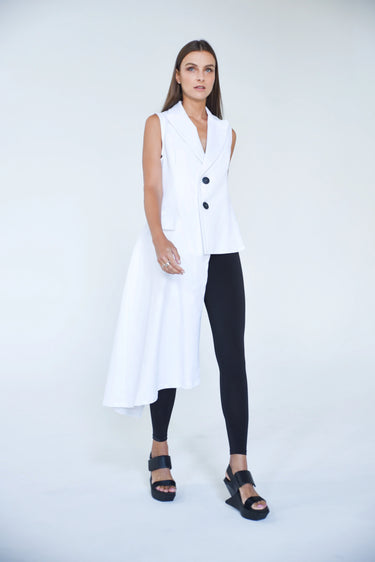 A woman standing against a white backdrop wearing an asymmetrical sleeveless White vest and Black leggings.