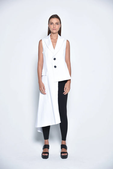 A woman standing against a white backdrop wearing an asymmetrical sleeveless White vest, Black leggings and black sandals.