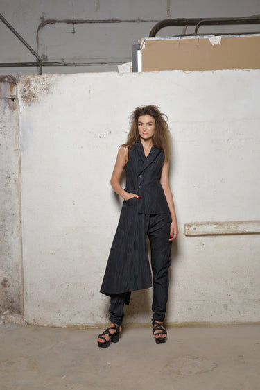 A woman standing against a white backdrop wearing an asymmetrical sleeveless black  vest and black pants.