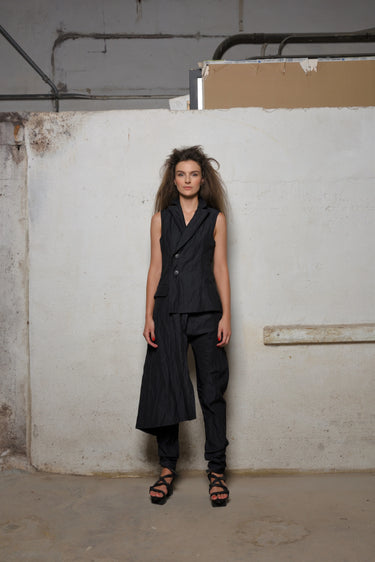 A woman standing against a white backdrop wearing an asymmetrical sleeveless black  vest and black pants.