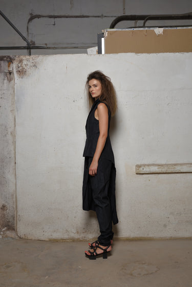 A woman standing against a white backdrop wearing an asymmetrical sleeveless black  vest and black pants.