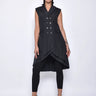 A woman wearing a sleeveless black trench-style Vest with a double-breasted button front, notched lapel collar and flap pockets on either side of the waist, asymmetrical hem line, paired with black leggings and chunky black open-toe heels over a white background.