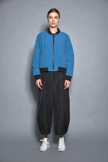 A woman wearing a blue bomber jacket and black pants against a gray background