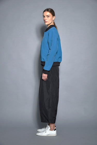 A woman wearing a blue bomber jacket and black pants against a gray background