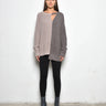 A woman standing against a white wall, wearing a 2 tone gray long-sleeve sweater with asymmetrical hem and black leggings.